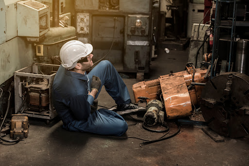 An injured industrial worker sits on the floor after an accident, surronded by machinery