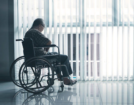 An elderly gentleman sits in a wheelchair looking out the window of a nursing home