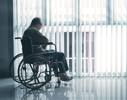 An elderly man looking sadly out a window at a nursing home.