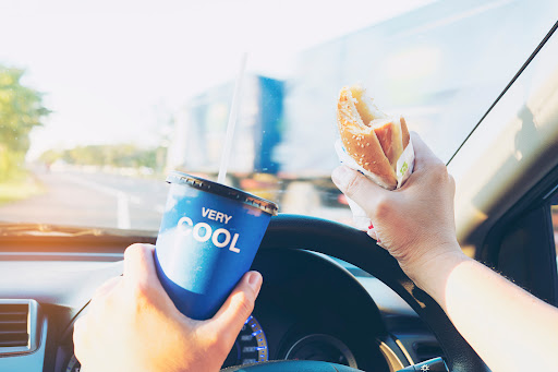 A person exhibits distracted driving by holding a hot dog in one hand and a drink in the other while driving in Louisiana.