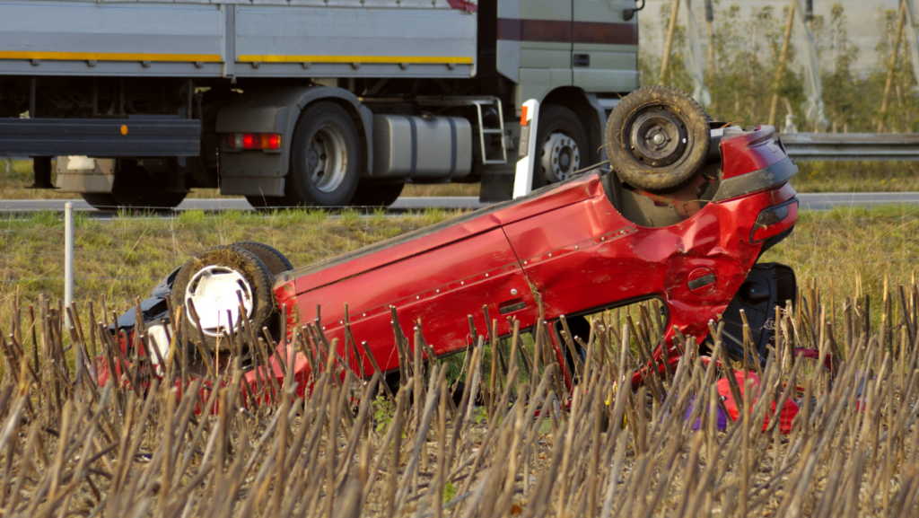 A car upside down in a field after an accident with the semi truck in the background
