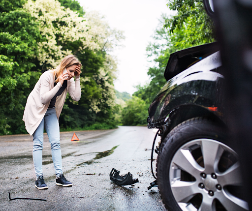 A distressed young woman on the phone surveying the damage to her car after an accident