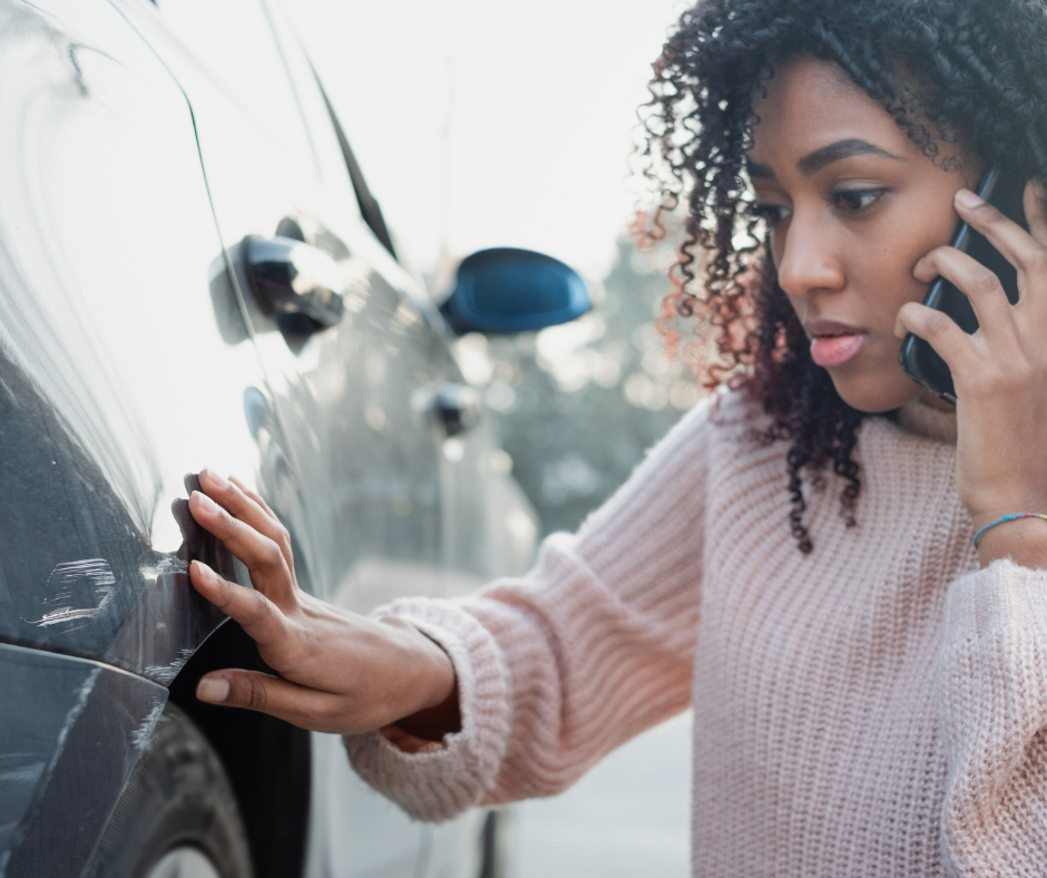 A young woman inspects damage to her car while talking on the phone