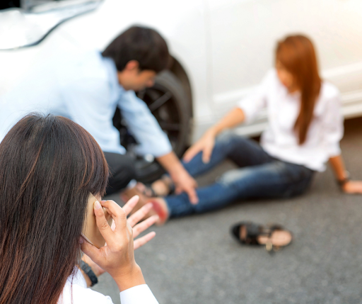 A man assisting with a woman's hurt ankle after a car accident while a brown-haired woman calls 911