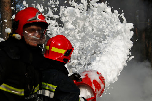 Two firefighters spray a large stream of Aqueous film forming foam, or AFF, from a firehose.