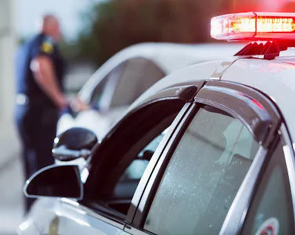 A closeup of a police car with glowing lights as an officer interacts with a driver in the background