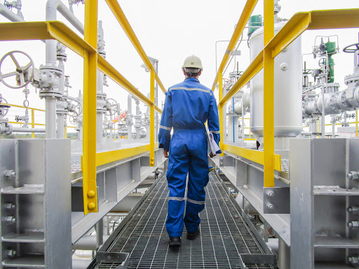 An industrial plant employee walks down a catwalk surrounded by pipes and machinery