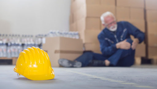 An injured warehouse worker sits on the floor holding his knee in pain with a yellow hard hat in the foreground