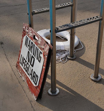 A sign that reads "Warning No Lifeguard" leaning against a lifeguard stand at an outdoor pool