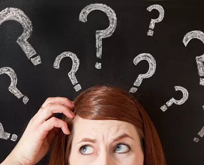 A woman scratching her head in confusion with an array of question marks on a chalkboard behind her