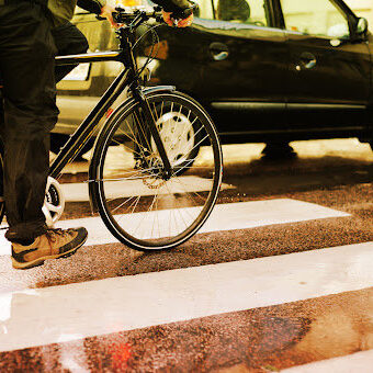Bicyclist in front of cars, wainting to cross road in rain and sun.