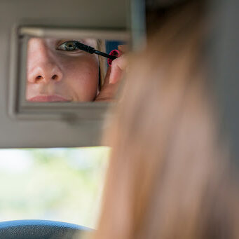 A Caucasian teenage girl is sitting in the driver's seat of her new black car. She is applying makeup with the mirror while driving. She is driving recklessly.